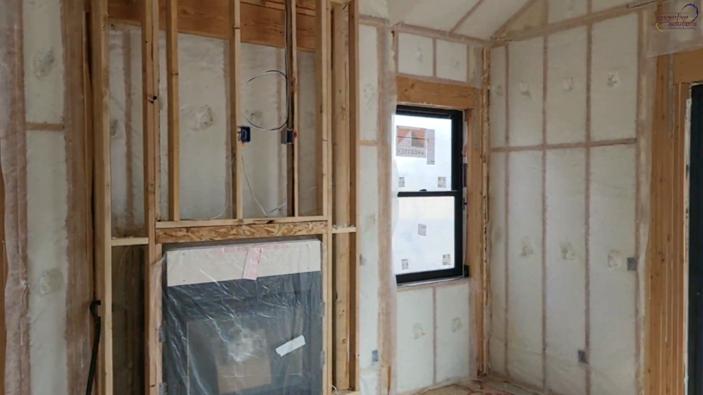 Interior wall framing under construction with exposed wooden studs, spray foam insulation applied between cavities, visible electrical wiring, a window opening letting in natural light, and an unfinished fireplace section prepared for drywall installation.