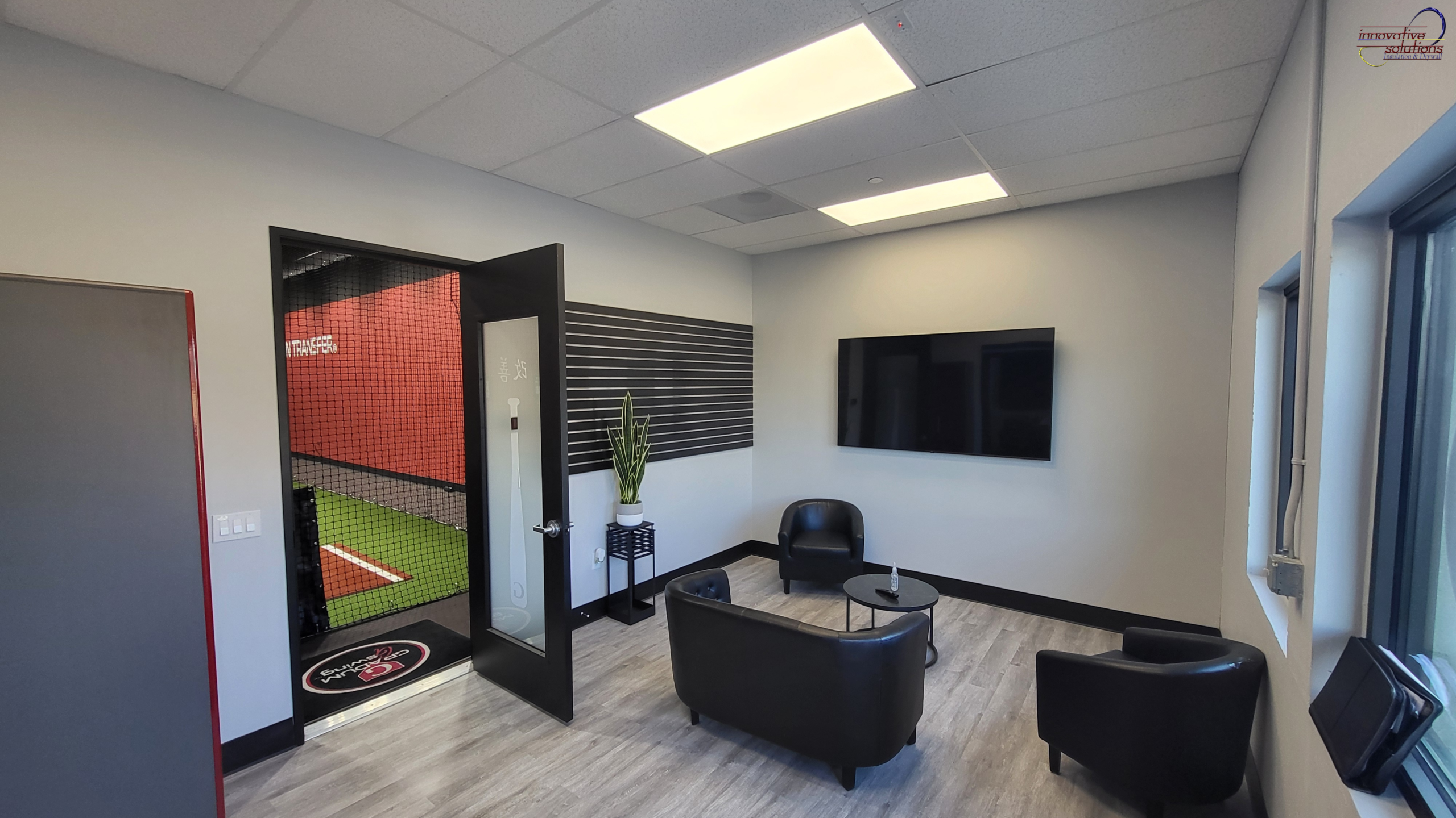 Modern finished interior space with smooth drywall walls and ceiling tiles, featuring a small seating area with black chairs and table, wall-mounted TV, indoor plant, and an open door leading to a sports training area, showcasing a clean and professional drywall finish.
