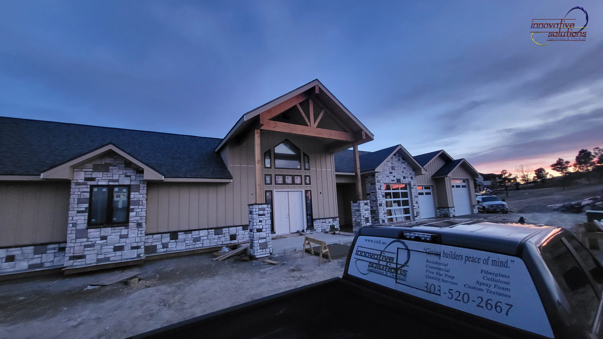 Modern custom luxury home exterior under construction at dusk with stone accents, large timber entryway, and contractor truck parked in front on a residential job site.