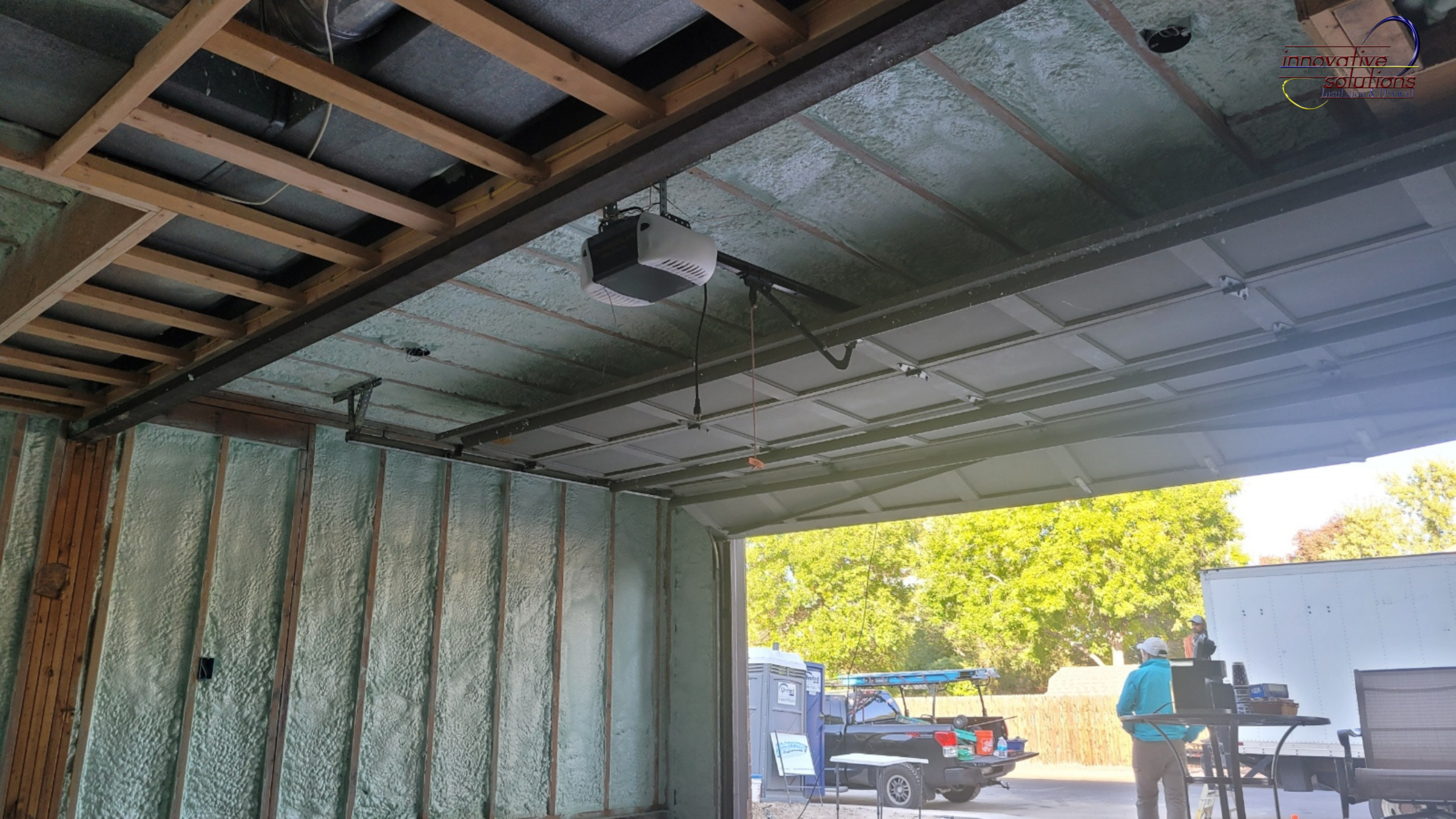 Interior view of a residential garage with spray foam insulation applied to walls and ceiling, exposed framing, open garage door, and workers outside during a home construction project.
